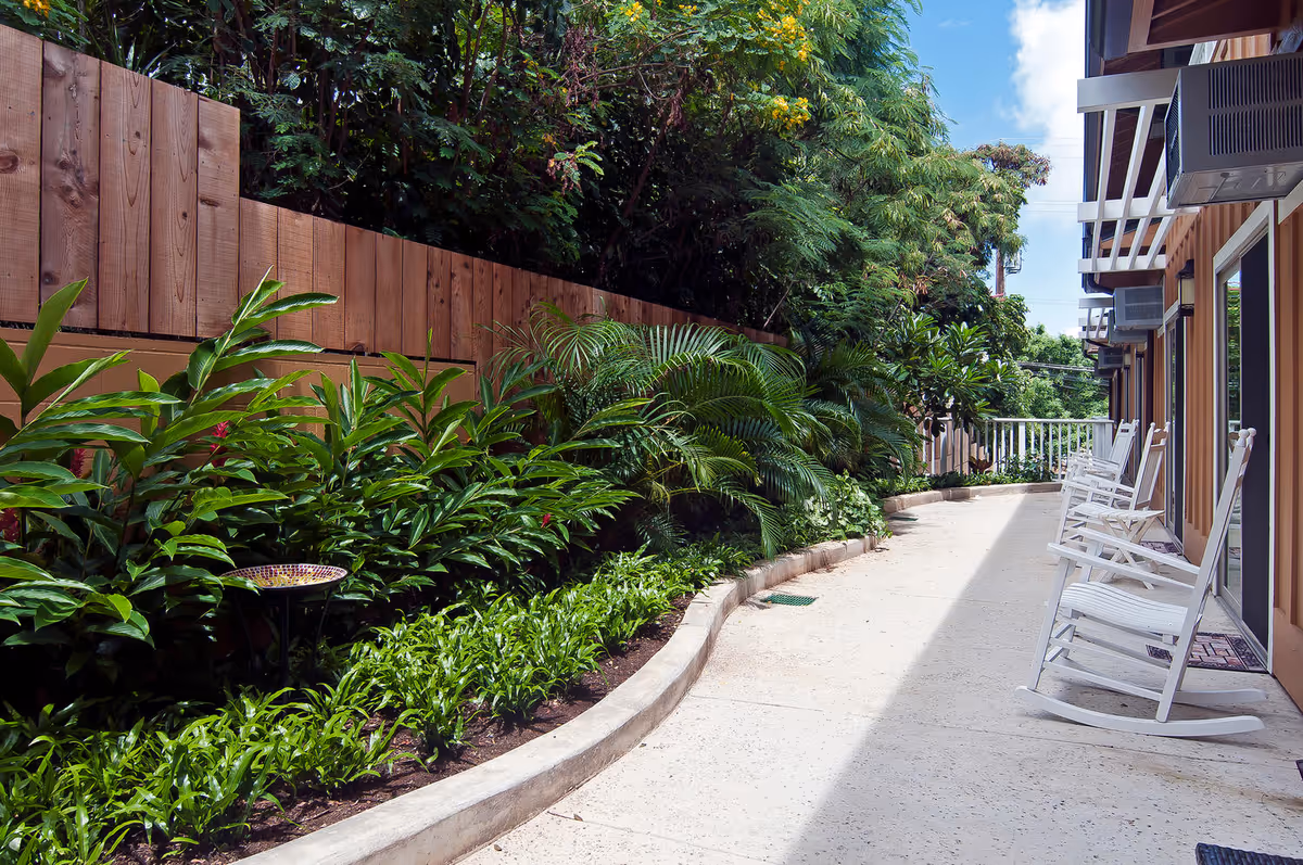 Outdoor patio area at Wilson Senior Living featuring a curved concrete walkway lined with lush green plants and a wooden fence on one side. On the other side, there are several white rocking chairs placed in front of building doors with air conditioning units above. The sky is partly cloudy and blue.