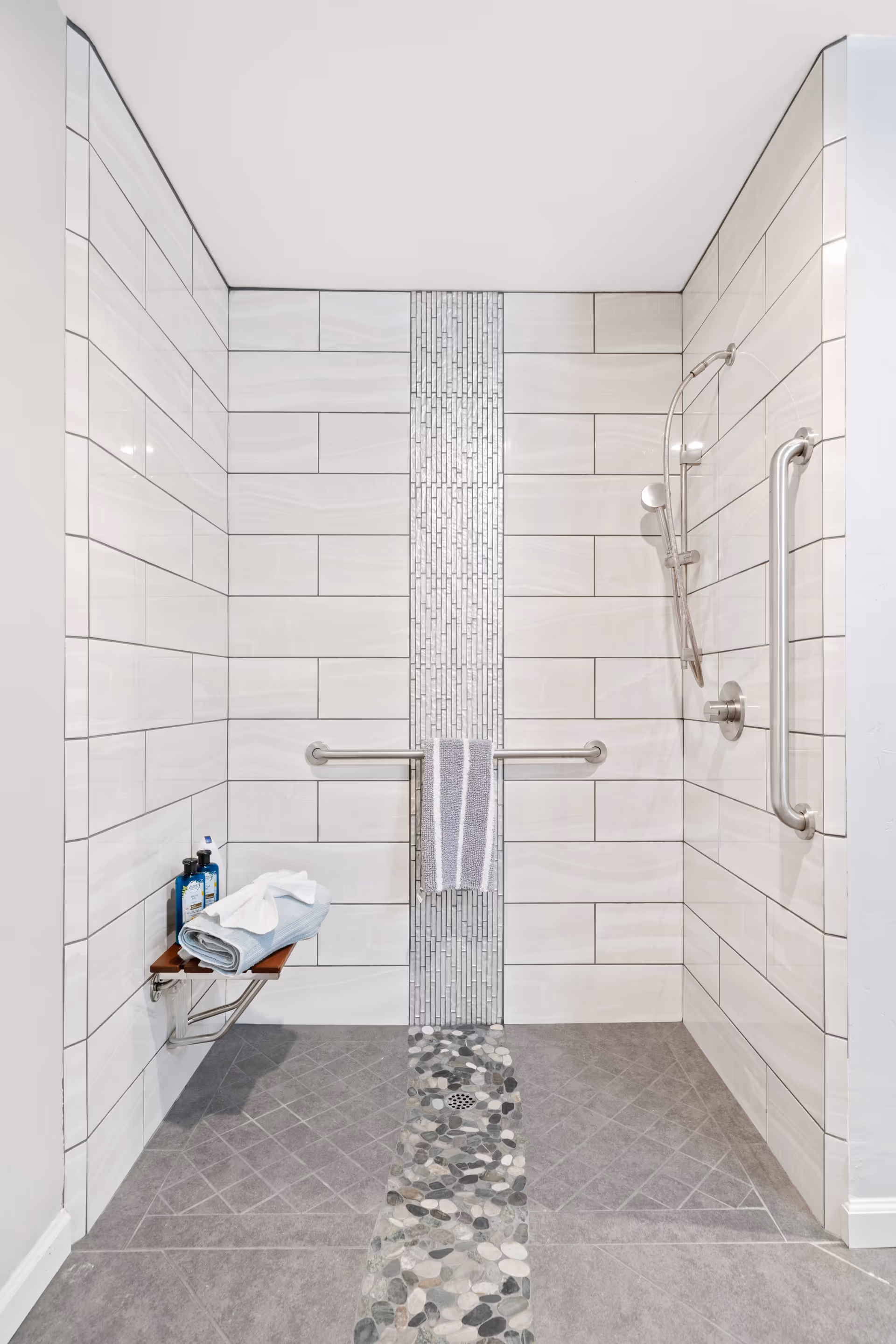 A modern walk-in shower with light gray rectangular tiles on the walls and a vertical strip of small mosaic tiles in the center. The floor has gray tiles with a pebble stone strip leading to the drain. There is a foldable wooden shower seat with towels and bottles on it, a horizontal grab bar with a towel hanging, and a vertical grab bar next to a handheld showerhead.