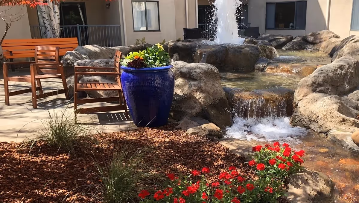 Outdoor seating area with wooden chairs and benches near a decorative water fountain surrounded by rocks, plants, and flowers in a garden setting at Mercy McMahon Terrace.