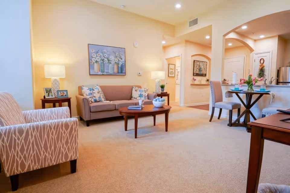 A cozy living room with beige walls and carpet, featuring a brown sofa with floral cushions, a patterned armchair, a wooden coffee table with a bowl of decorative balls and a book, two side tables with lamps and framed photos, and a small dining area with a round table set for two. The room opens into a kitchen area with stainless steel appliances and a hallway with artwork on the walls.