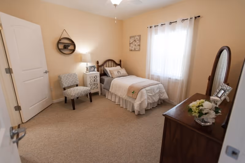 A cozy bedroom in a senior living facility featuring a single bed with white and beige bedding, a wooden headboard, a small white nightstand with a lamp and picture frame, a patterned chair, a wooden dresser with a mirror and flower arrangement, beige carpet, cream-colored walls, and a window with sheer white curtains.