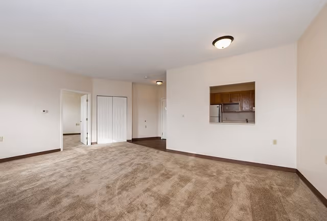 Empty living room with beige carpet and white walls, featuring a small pass-through window to a kitchen with wooden cabinets and a refrigerator. There is a doorway leading to another room and a closet with white double doors.