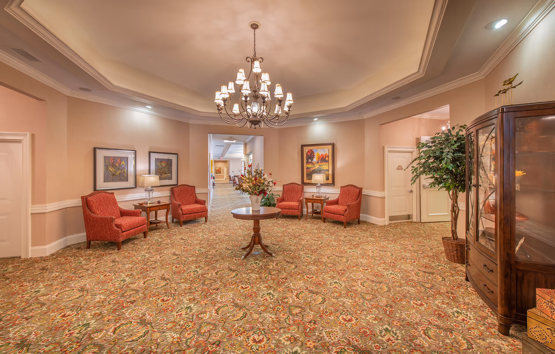 A spacious and elegantly decorated common area with a patterned carpet, four red upholstered armchairs arranged in pairs on either side of the room, two small wooden side tables with lamps, a round wooden table with a floral arrangement in the center, a large chandelier hanging from the ceiling, framed artwork on the walls, a tall potted plant, and a wooden display cabinet on the right side.