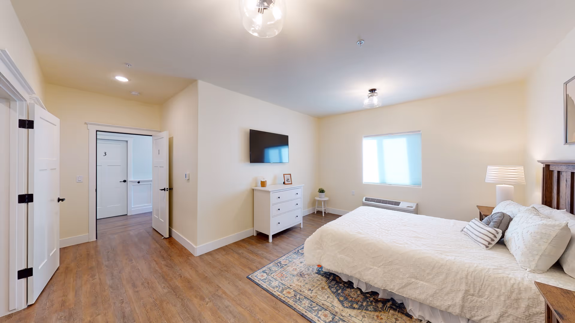 A bright and clean bedroom in an assisted living facility featuring a large bed with white bedding and multiple pillows, a wooden headboard, a patterned area rug, a white dresser with decorative items, a wall-mounted flat screen TV, a window with a shade, and wooden flooring. The room has light-colored walls and ceiling lights, with open doors leading to a hallway.