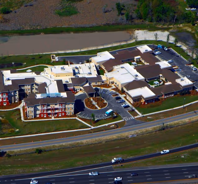 Aerial view of Promise Pointe Senior Living facility showing multiple connected buildings with white and brown roofs, parking lots with several cars, surrounding green lawns, a nearby road, and a body of water in the background.