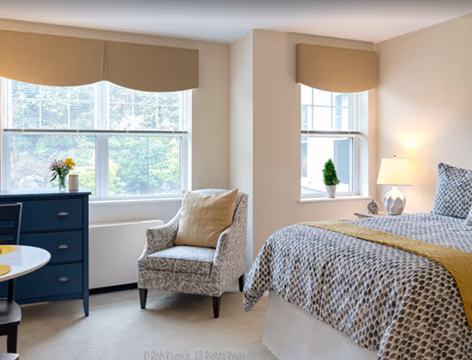 Bright, neatly furnished bedroom with a bed, patterned armchair, blue dresser and two windows letting in daylight.