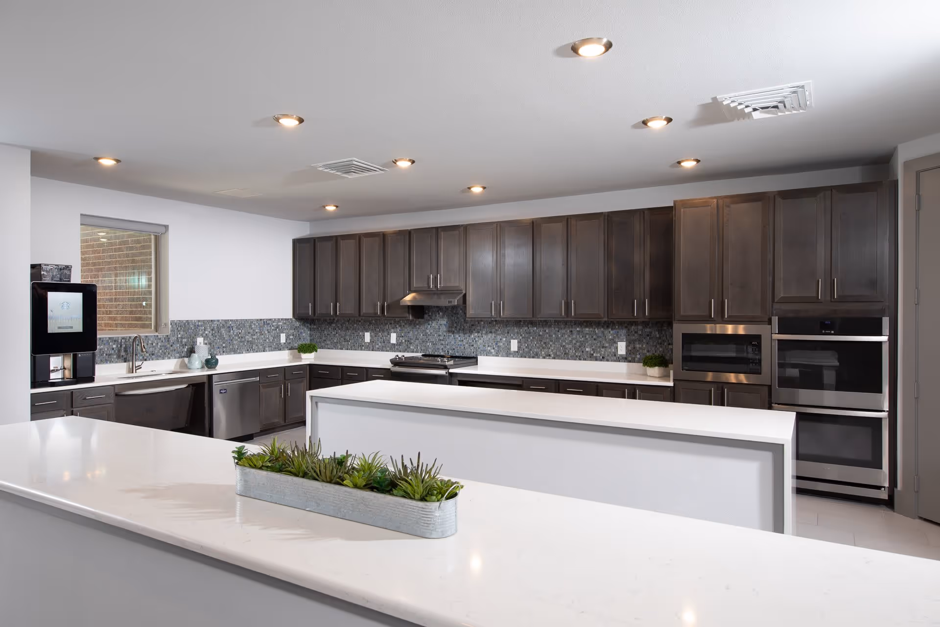 Modern kitchen with dark wood cabinets, white countertops, a mosaic tile backsplash, built-in microwave and oven, a stove with a hood, a dishwasher, a coffee machine, and a rectangular planter with green plants on the counter.