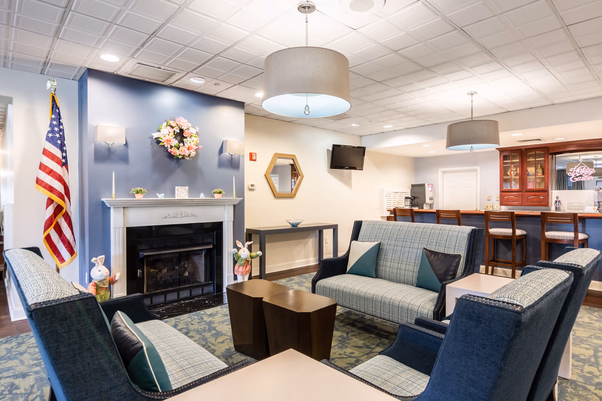 A cozy living room area in a senior living facility with blue and plaid upholstered sofas and armchairs arranged around a dark wooden coffee table. The room features a white fireplace with a floral wreath above it, an American flag to the left, and two decorative bunny figurines on either side of the fireplace. There is a blue accent wall behind the fireplace, a hexagonal mirror on the adjacent wall, a small table beneath it, and a wall-mounted TV. In the background, there is a bar area with wooden bar stools and a neon sign that reads 'Family Center'. The ceiling has recessed lighting and two large pendant lights.