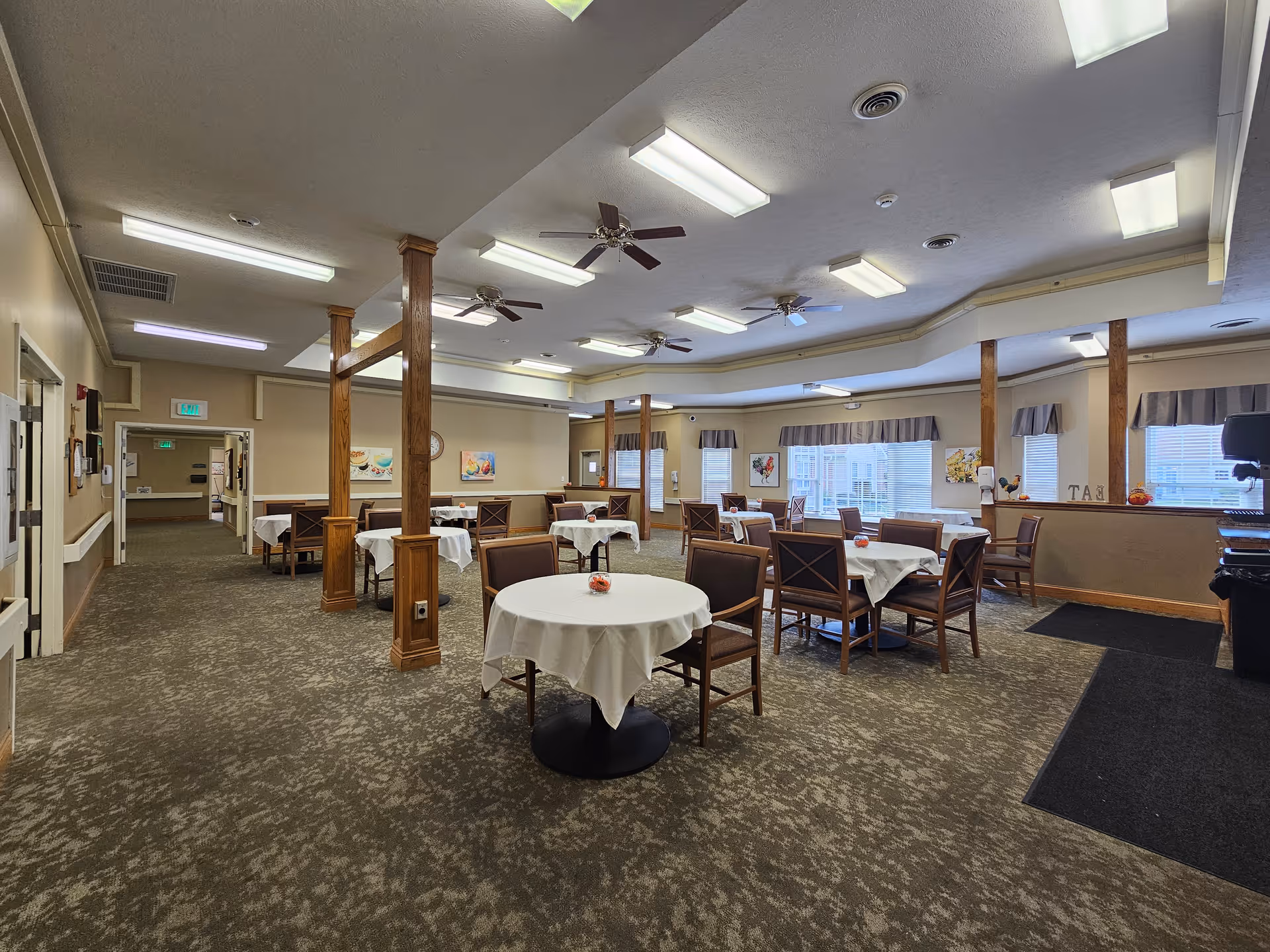 A spacious dining room with multiple round tables covered with white tablecloths, each surrounded by wooden chairs with cushions. The room has beige walls, carpeted floors, several ceiling fans, and fluorescent lighting. Windows with valances let in natural light, and there are decorative paintings on the walls. Wooden pillars divide the space, and there is a black mat near the entrance area.