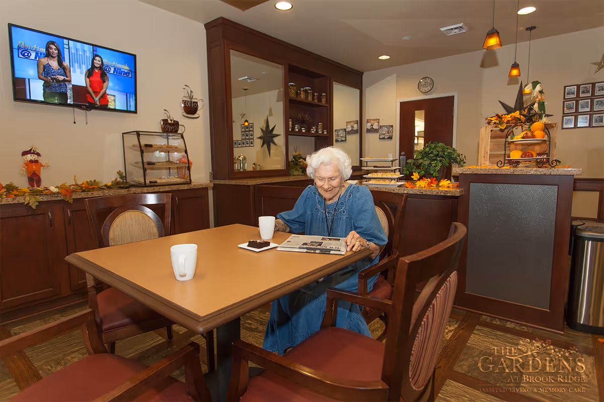 An elderly woman sitting at a dining table in a cozy room, reading a newspaper with a cup of coffee and a brownie on a plate in front of her. The room has wooden cabinets, a TV mounted on the wall showing two women on a news program, and autumn-themed decorations. There is a fruit basket and plants on a counter in the background.