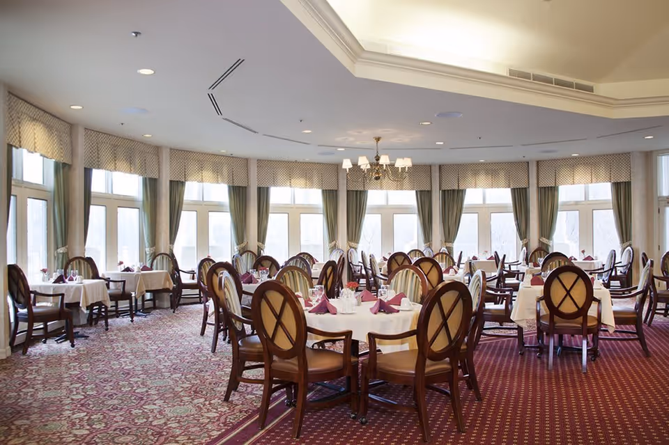 A spacious dining room with multiple round tables covered with white tablecloths and set with folded burgundy napkins, glassware, and silverware. The room features large windows with green curtains and valances, a patterned carpet, and a chandelier hanging from the ceiling.