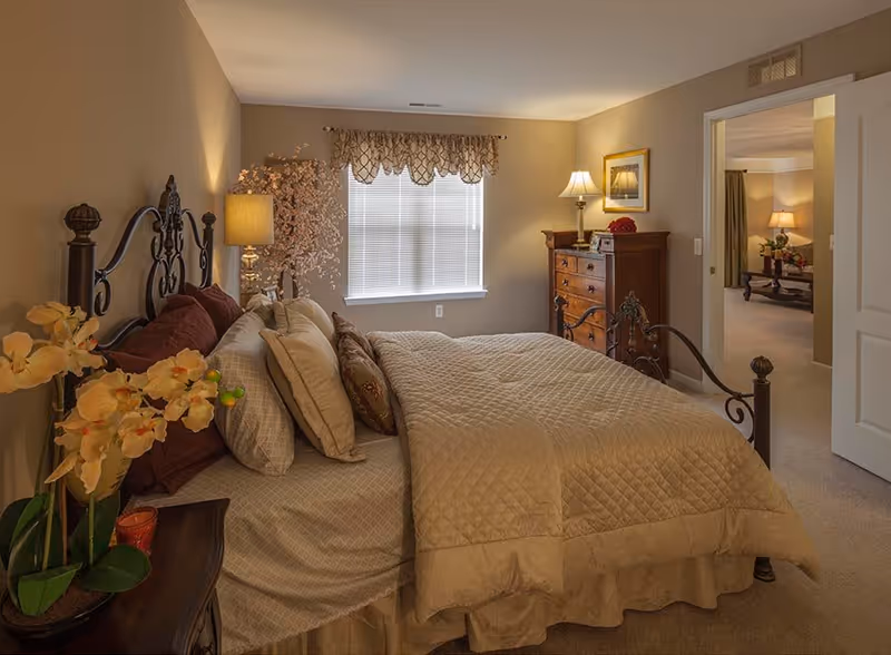 A warmly lit bedroom with a large bed covered in beige quilted bedding and multiple pillows. The bed has an ornate dark metal headboard. There is a wooden nightstand with a potted orchid and a lamp with a beige shade. A window with blinds and a decorative valance is centered on the far wall. To the right, there is a wooden dresser with a lamp and framed artwork above it. An open door leads to another room with a lamp and furniture visible.