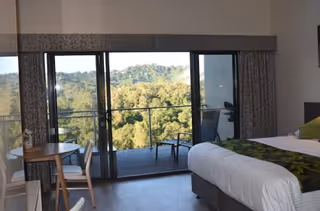 A bright bedroom featuring a bed, a small table with chairs, and sliding glass doors leading to a balcony with a wooded hill view.