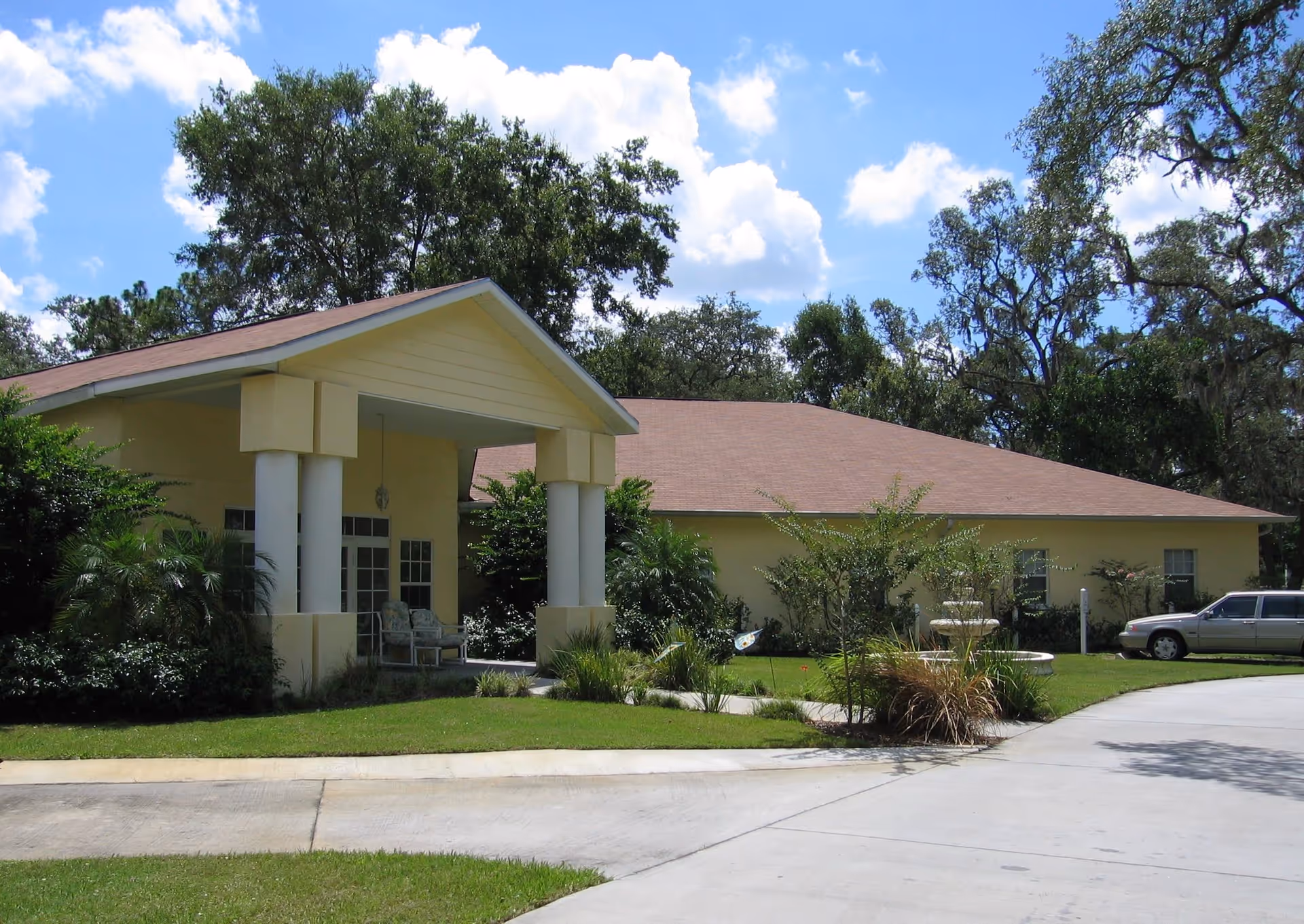 Front view of a single-story yellow building with a covered columned entrance, landscaped lawn and a curved driveway with a parked car.