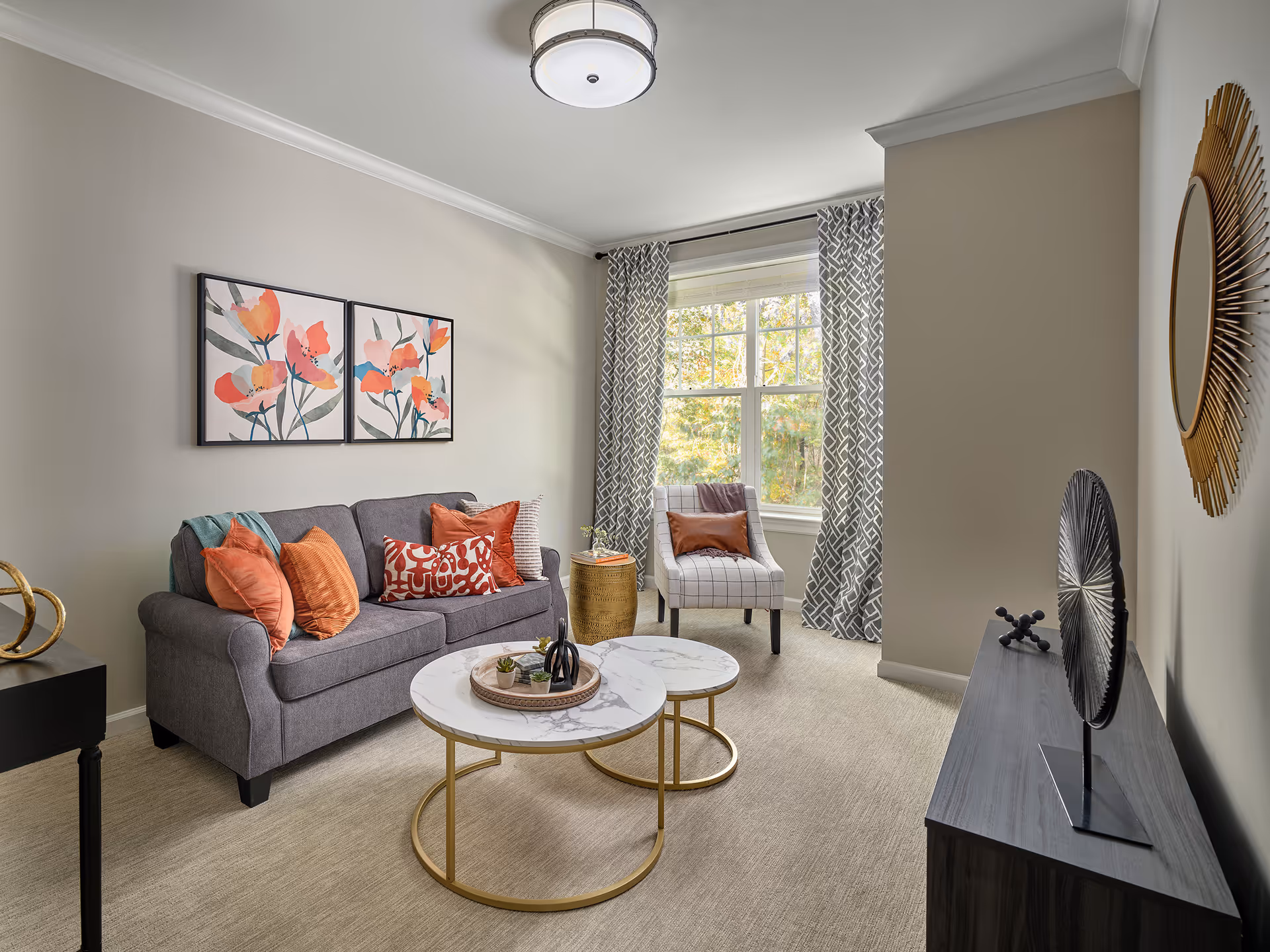 A cozy living room with a gray sofa adorned with orange and patterned throw pillows, a pair of round nested coffee tables with marble tops and gold legs, a patterned armchair with an orange pillow near a window with gray and white curtains, and decorative wall art including floral paintings above the sofa and a sunburst mirror on the adjacent wall.