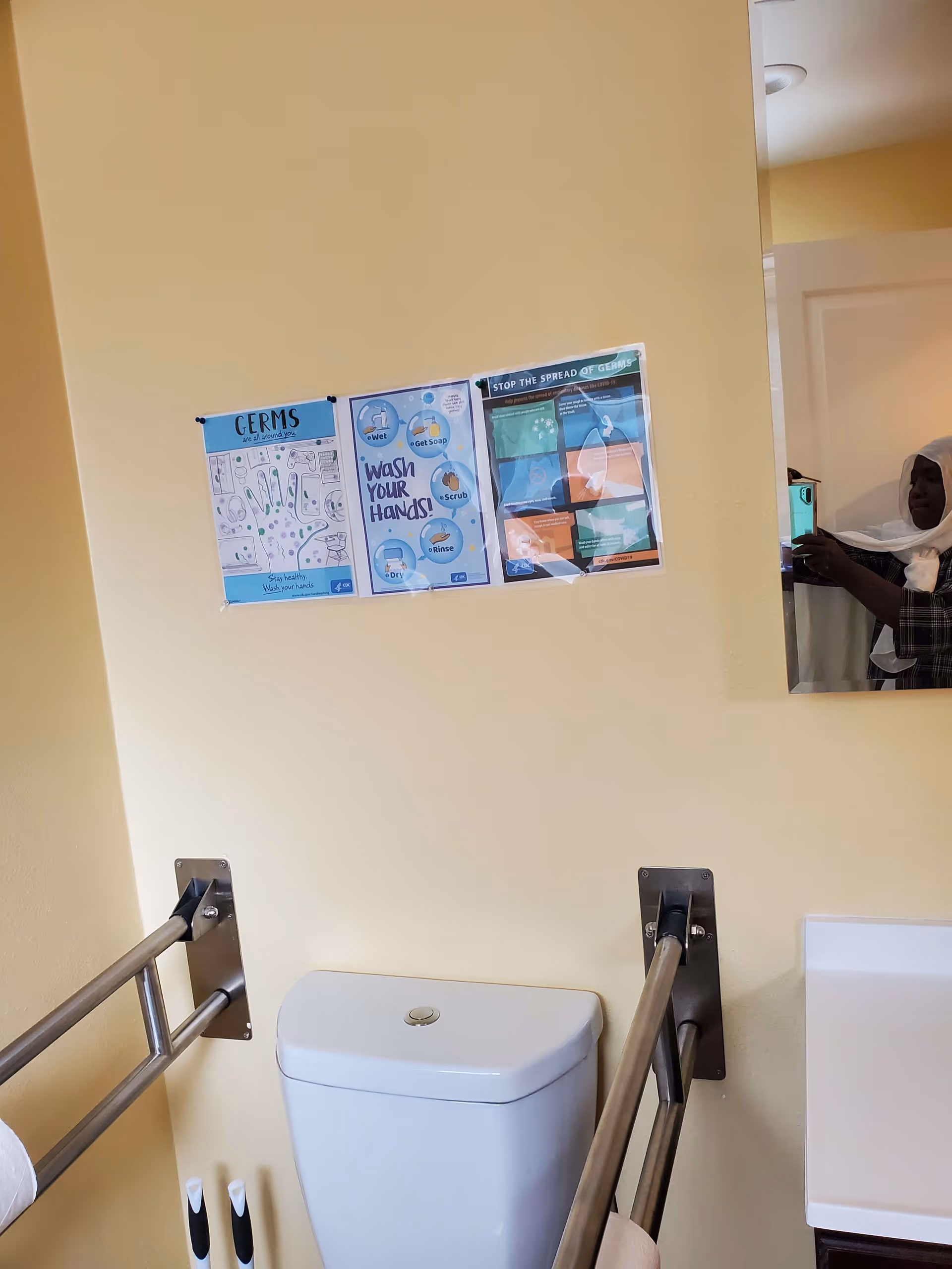 Accessible bathroom with a toilet, metal grab bars, handwashing posters on a pale yellow wall, and a mirror reflecting the person taking the photo.