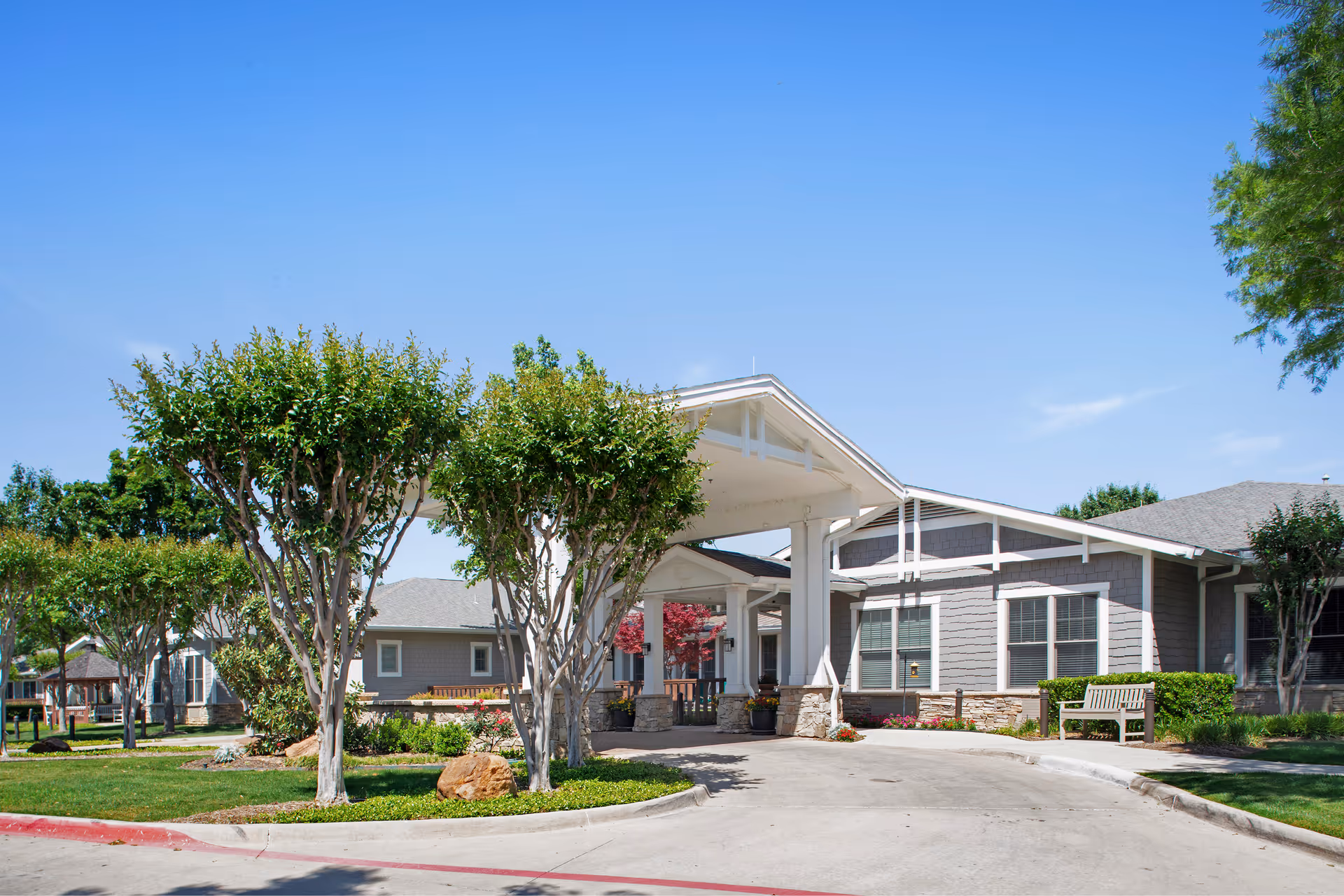 Front entrance of Sunrise of Fort Worth with a covered porte-cochère, landscaped trees, and benches.