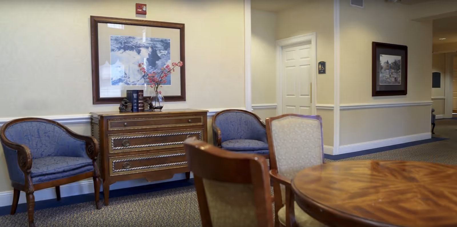 A furnished senior living lounge with a round wooden table and chairs, two upholstered armchairs, a decorative chest with a vase and books, and framed artwork on the walls.
