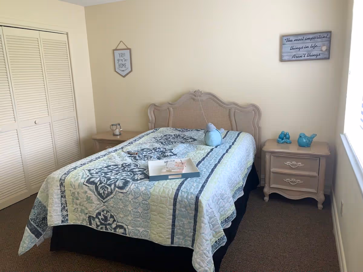 Light, simply furnished bedroom with a patterned quilted bed, two bedside tables, decorative items and a louvered closet door.
