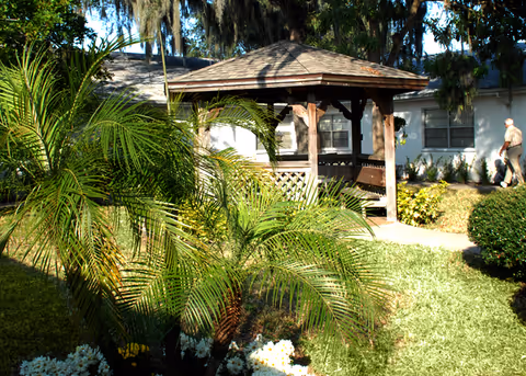 Wooden gazebo surrounded by palm plants and lawns in a courtyard with a light-colored building and a person walking in the background.