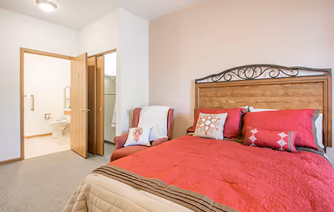 A bedroom with a wooden bed frame and red bedding, including decorative pillows. To the left, an open door reveals a bathroom with a toilet and grab bars, and a closet with hanging clothes. There is also a cushioned armchair with a white throw and a pillow next to the bed.