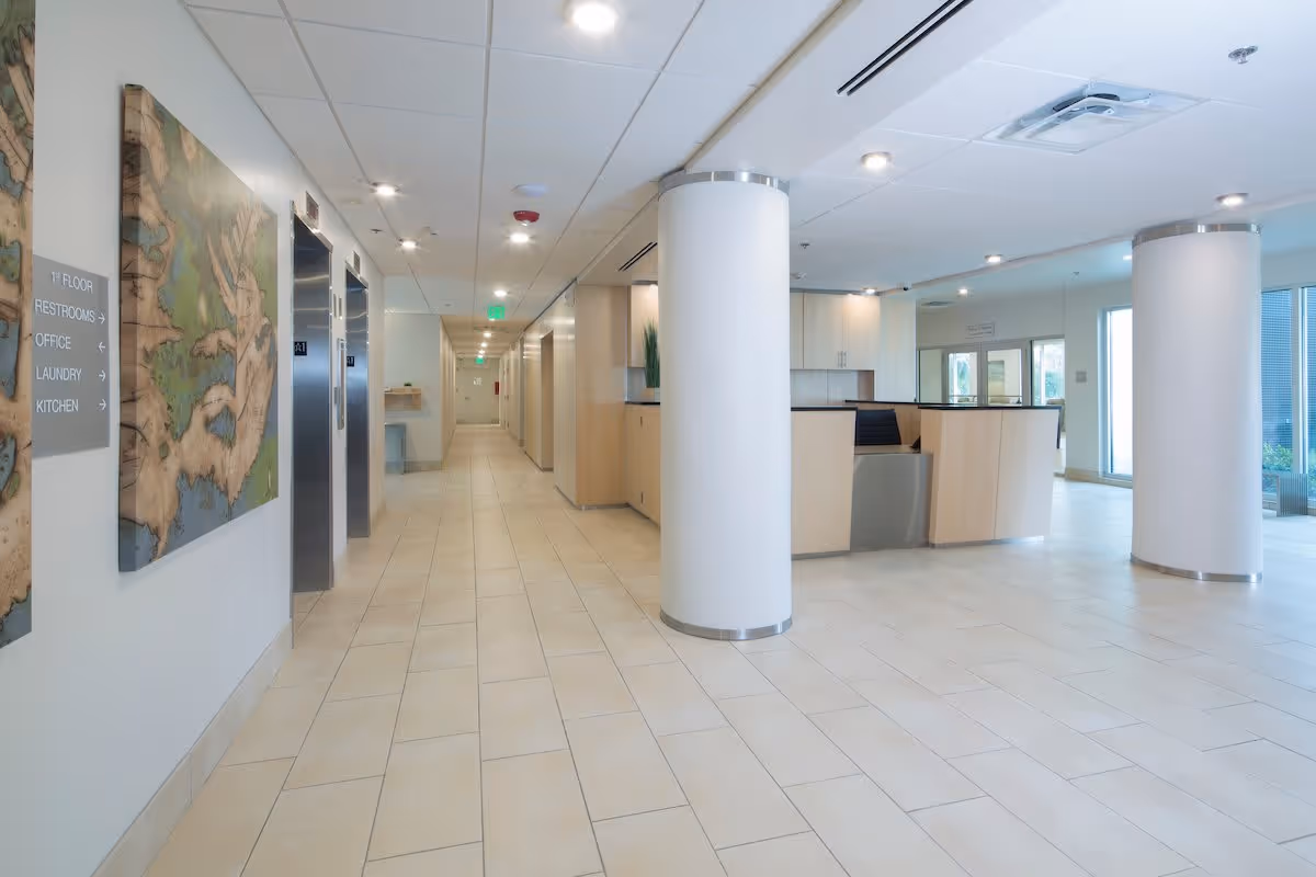 Bright modern lobby and long hallway with a reception desk, white columns, elevators and directional signage.
