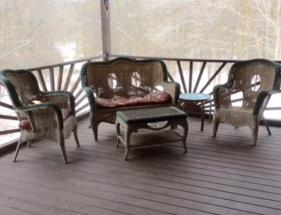 Wicker porch furniture set with two chairs, a loveseat, a coffee table and a side table on a covered wooden deck overlooking a snowy wooded area.