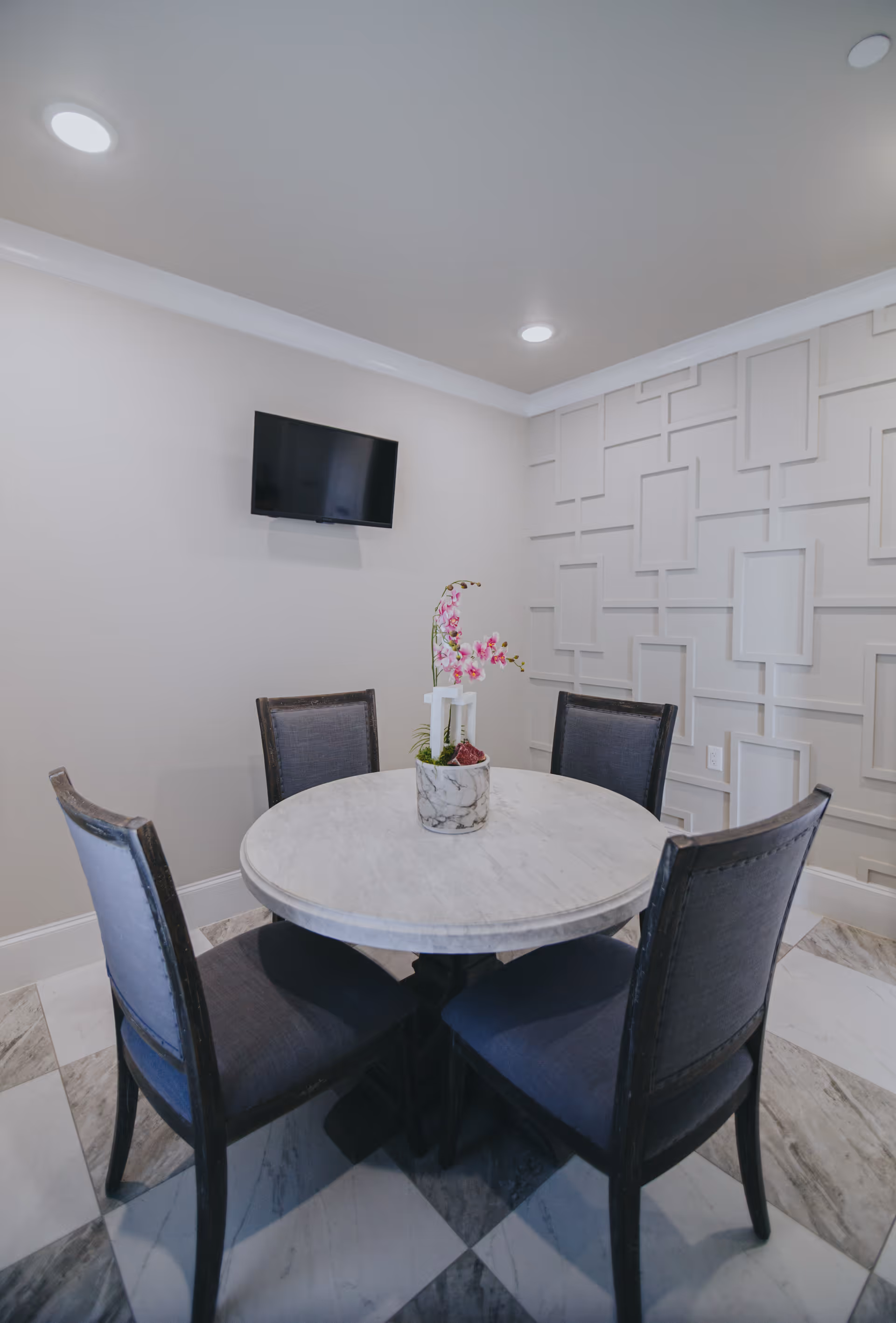 Small round marble table with four upholstered chairs and a floral centerpiece in a modern dining nook with a wall-mounted TV and decorative paneled wall.