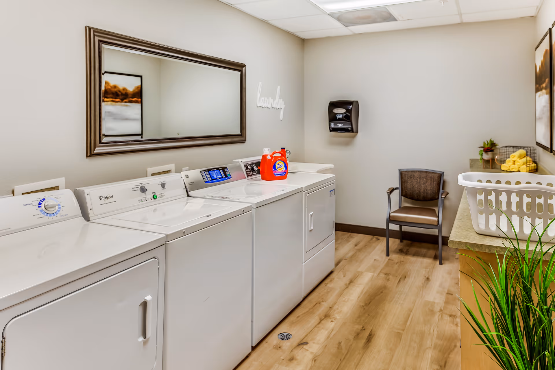 Laundry room with three white washing machines and dryers lined up against a beige wall. A large rectangular mirror hangs above the machines, and a bottle of Tide detergent is placed on one of the machines. On the right side, there is a countertop with a white laundry basket, a small plant, and neatly rolled yellow towels. A brown chair is positioned against the far wall beneath a paper towel dispenser. The floor is wooden, and the walls are painted light beige.