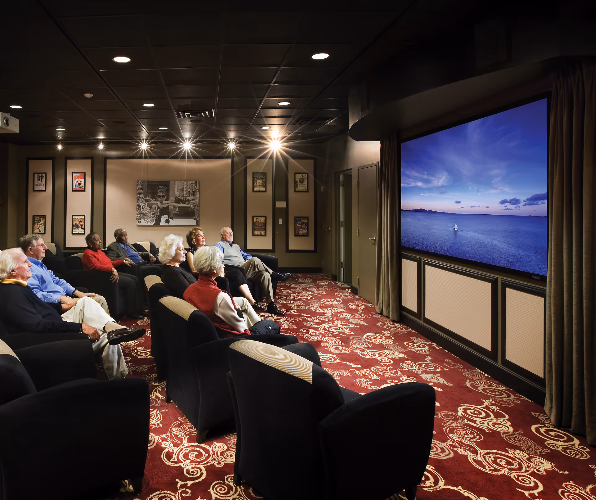 A group of older adults seated in a cozy screening room watching a large wall-mounted TV showing a calm ocean scene.