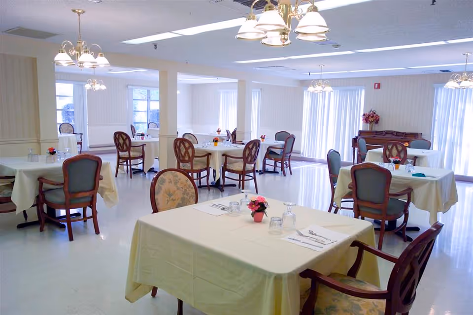 A bright dining room with multiple tables covered in cream-colored tablecloths, each set with glasses, napkins, and small flower arrangements. The room features wooden chairs with cushioned seats and backs, chandeliers hanging from the ceiling, large windows with vertical blinds, and a piano against the far wall.