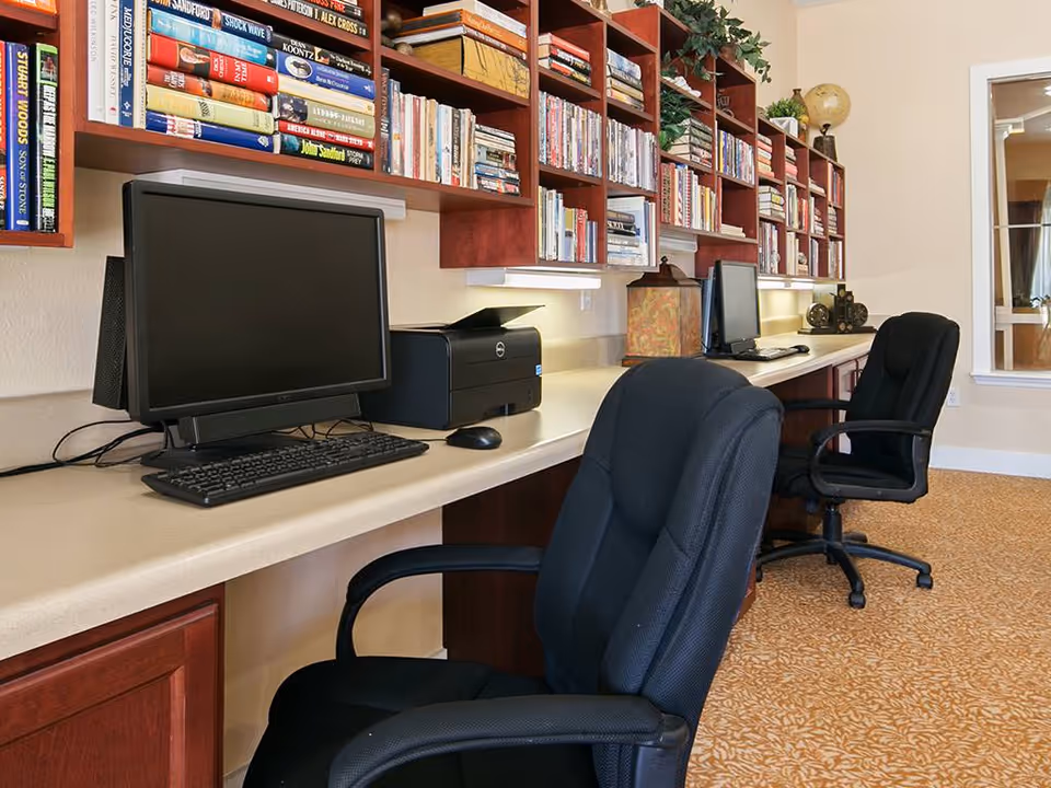 A computer workstation area with two black office chairs, two desktop computers, a printer, and a long countertop. Above the countertop are wooden shelves filled with books and decorative items. The room has beige walls and patterned carpet flooring.