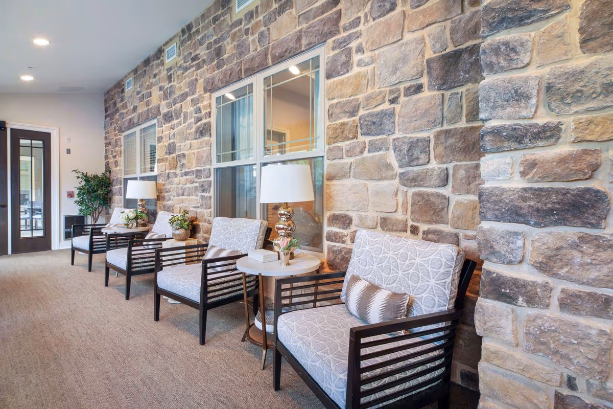 A cozy seating area with four cushioned armchairs arranged along a stone wall. Each chair has a small pillow and is paired with a round side table holding a decorative lamp and small plants. The space is well-lit with recessed ceiling lights and has a carpeted floor. A door with glass panels is visible at the end of the hallway.