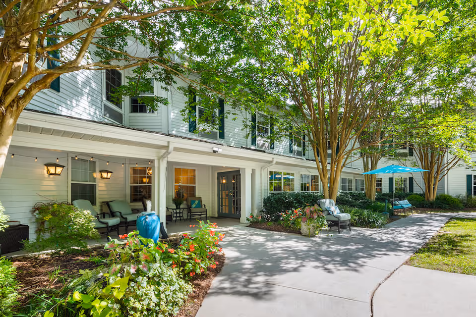 Outdoor patio area of Arbor Terrace Asheville with seating arrangements including cushioned chairs and a bench under a blue umbrella, surrounded by trees and landscaped flower beds in front of a white building with green shutters.