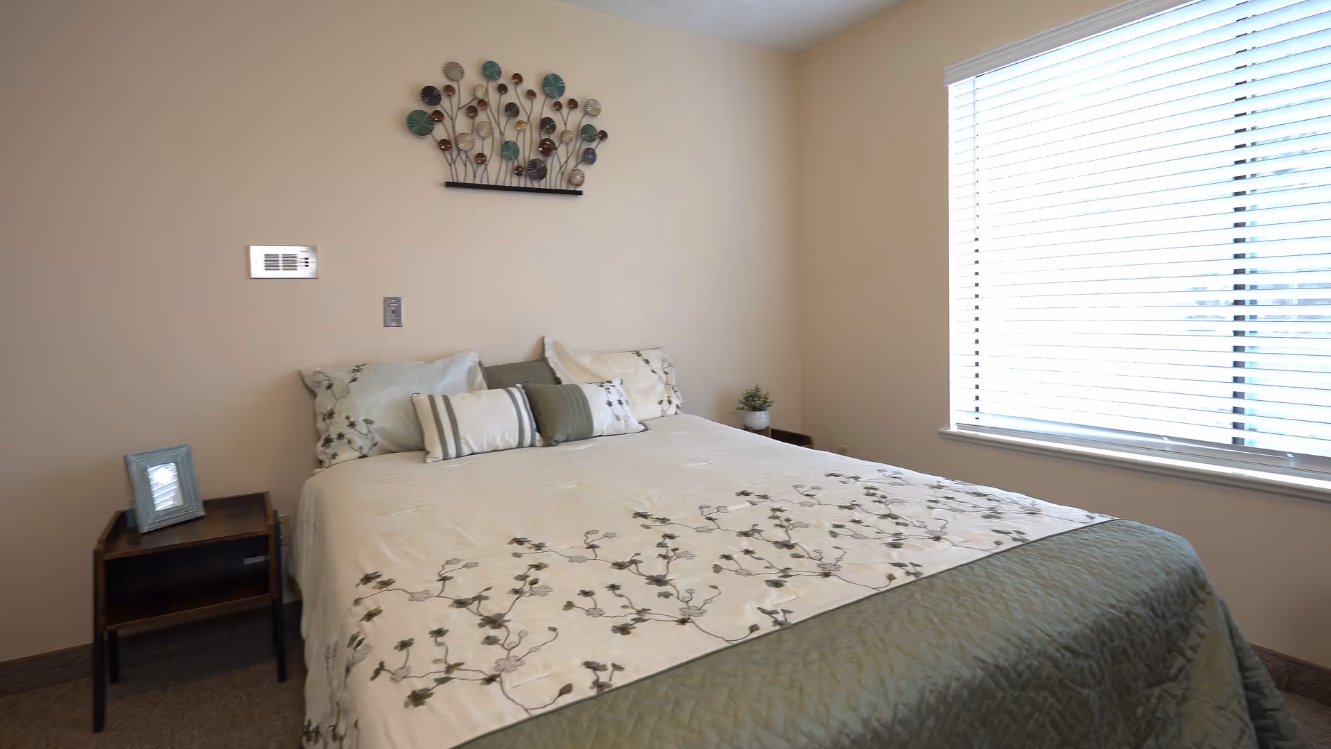 Sunlit bedroom with a made bed, decorative pillows, small nightstands and a large window with blinds.