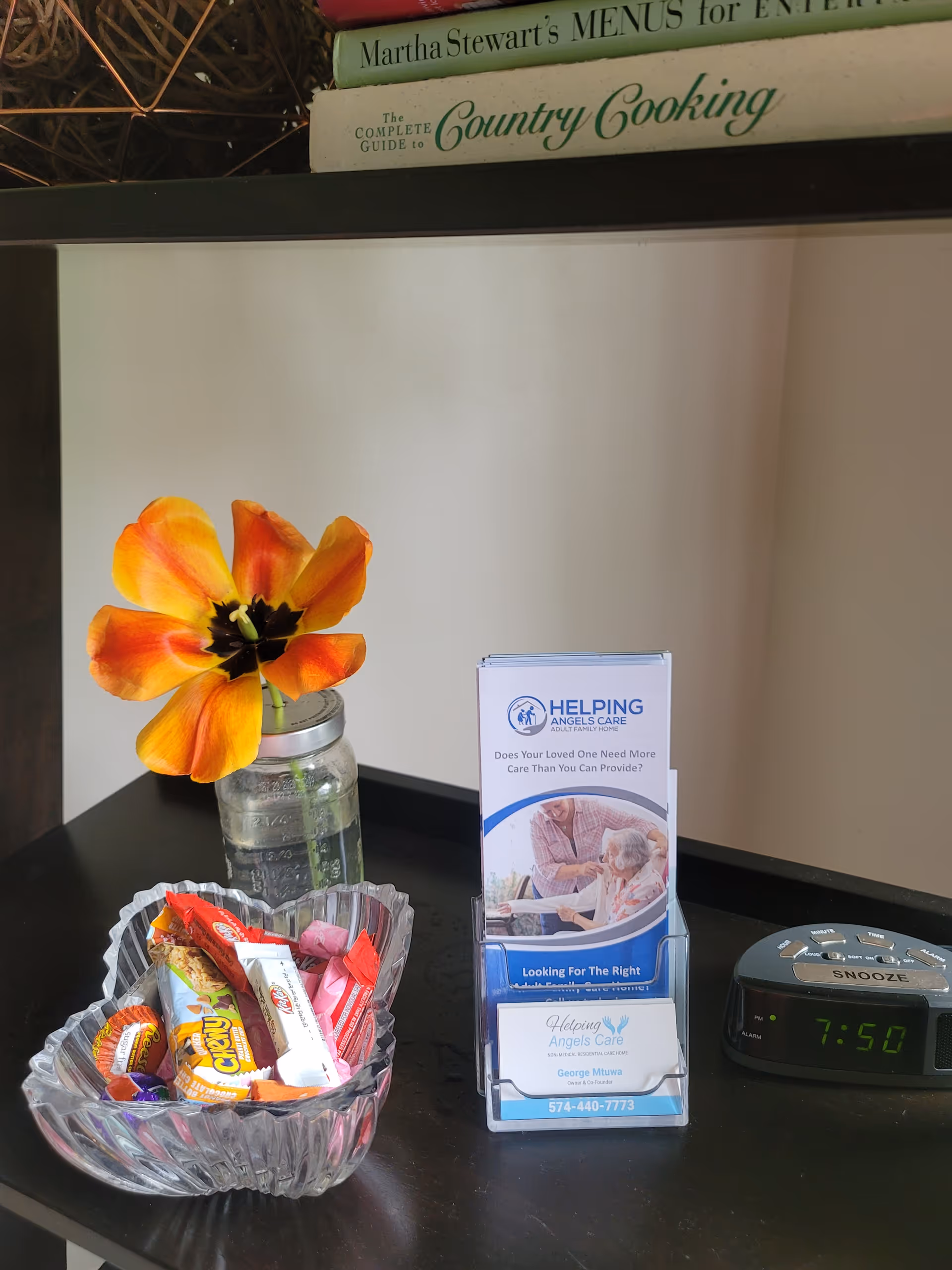 A small table with a mason jar holding an orange flower, a glass bowl of candy, a Helping Angels Care brochure display, and a digital alarm clock.