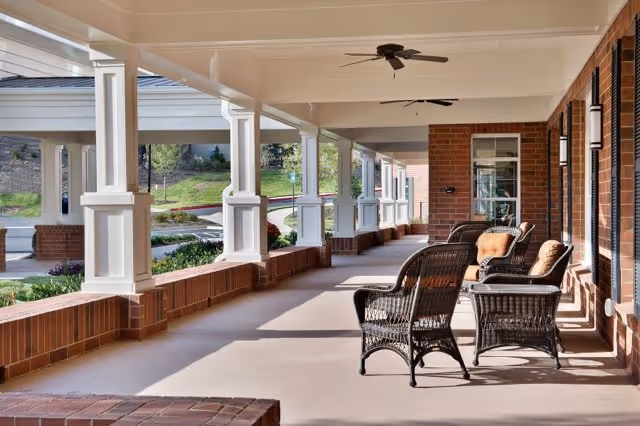 Covered outdoor patio area with wicker chairs and cushions arranged for seating. The patio has white columns, ceiling fans, and overlooks a landscaped garden area with grass and shrubs.