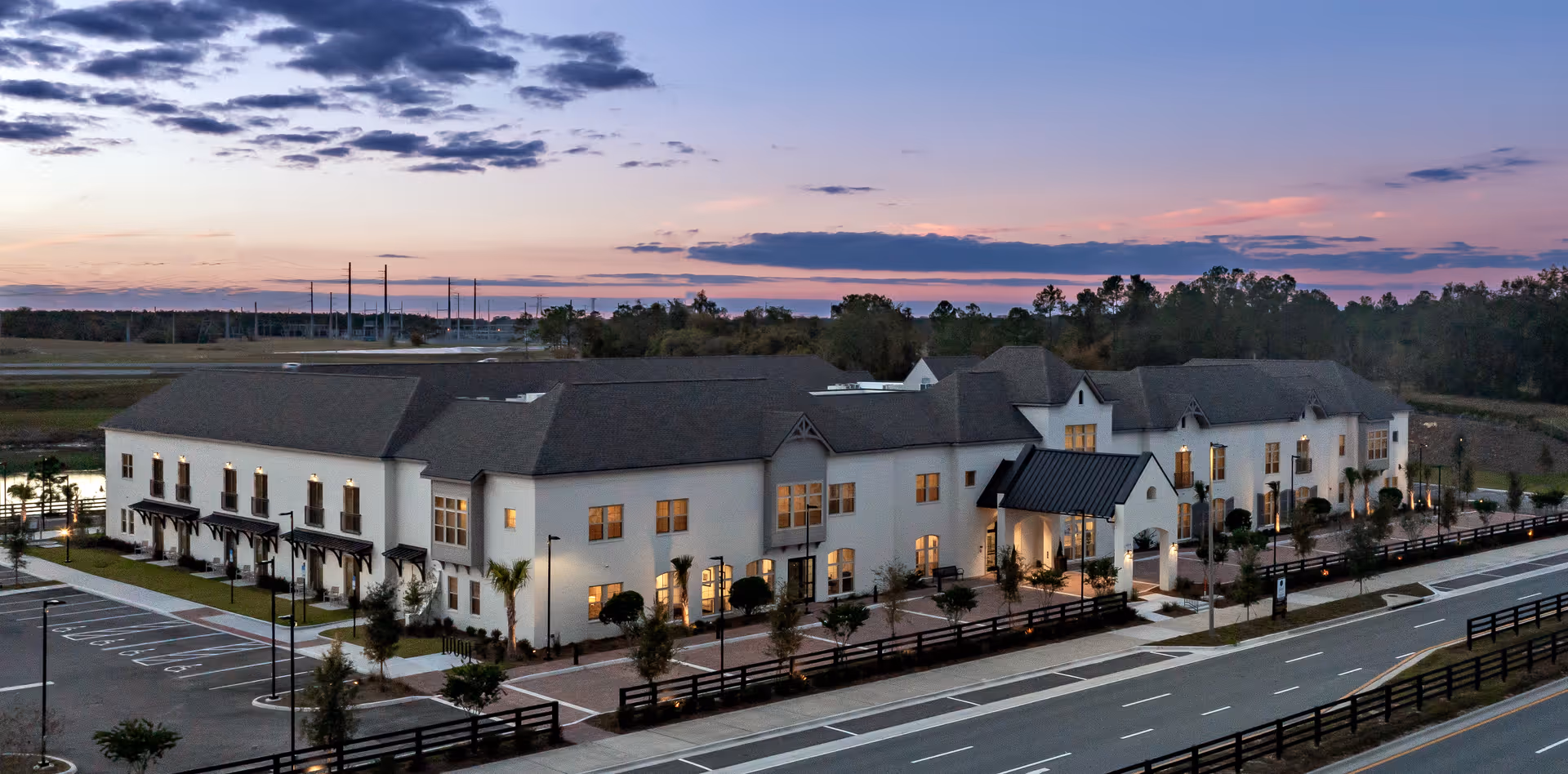 A large, two-story senior living facility building at dusk with lights on inside. The building has a modern design with a dark roof and white exterior walls. There is a paved road and parking area in front, with landscaped greenery and small trees around the property. The sky is partly cloudy with a colorful sunset in the background.