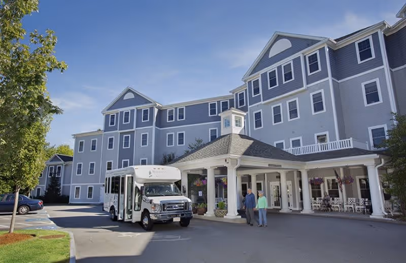 Exterior view of a multi-story senior living facility with gray siding and white trim. A white shuttle bus is parked in front of the covered entrance where two people are standing and talking. There are hanging flower baskets and outdoor seating on the porch area. Trees and a blue sky are visible in the background.