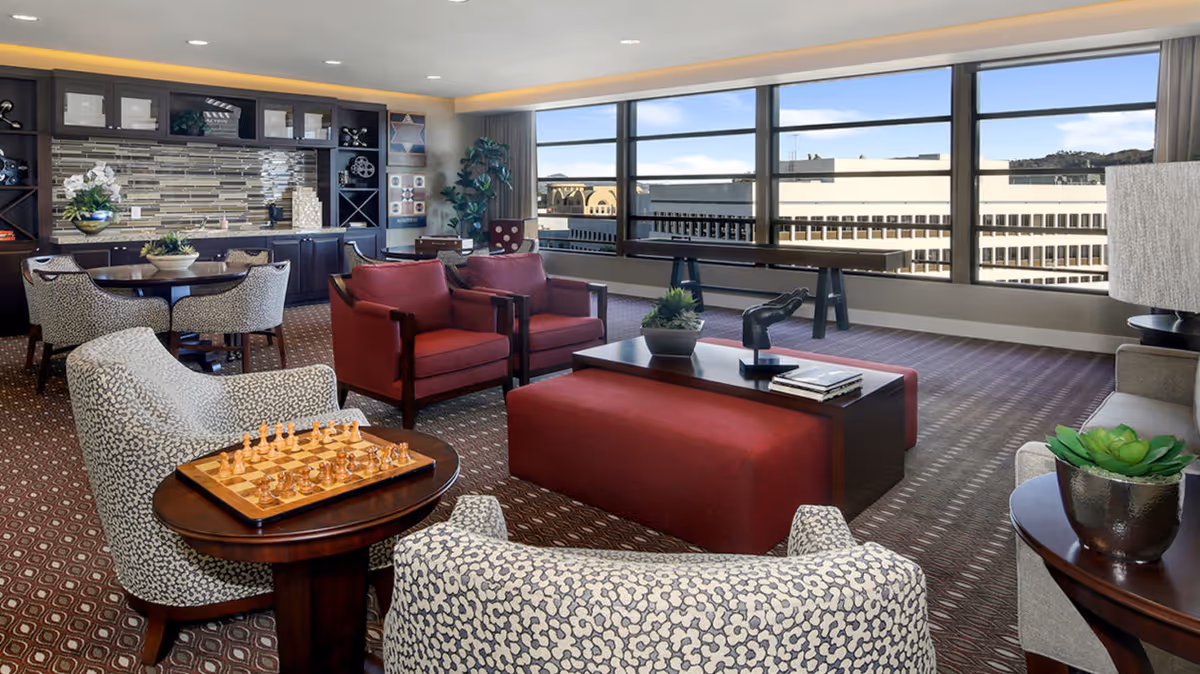 A spacious and well-lit common area with large windows offering a city view. The room features patterned armchairs, red upholstered chairs, a coffee table with decorative items, a chessboard on a side table, and a dining table with chairs. There is a built-in cabinet with shelves and decorative items against one wall.