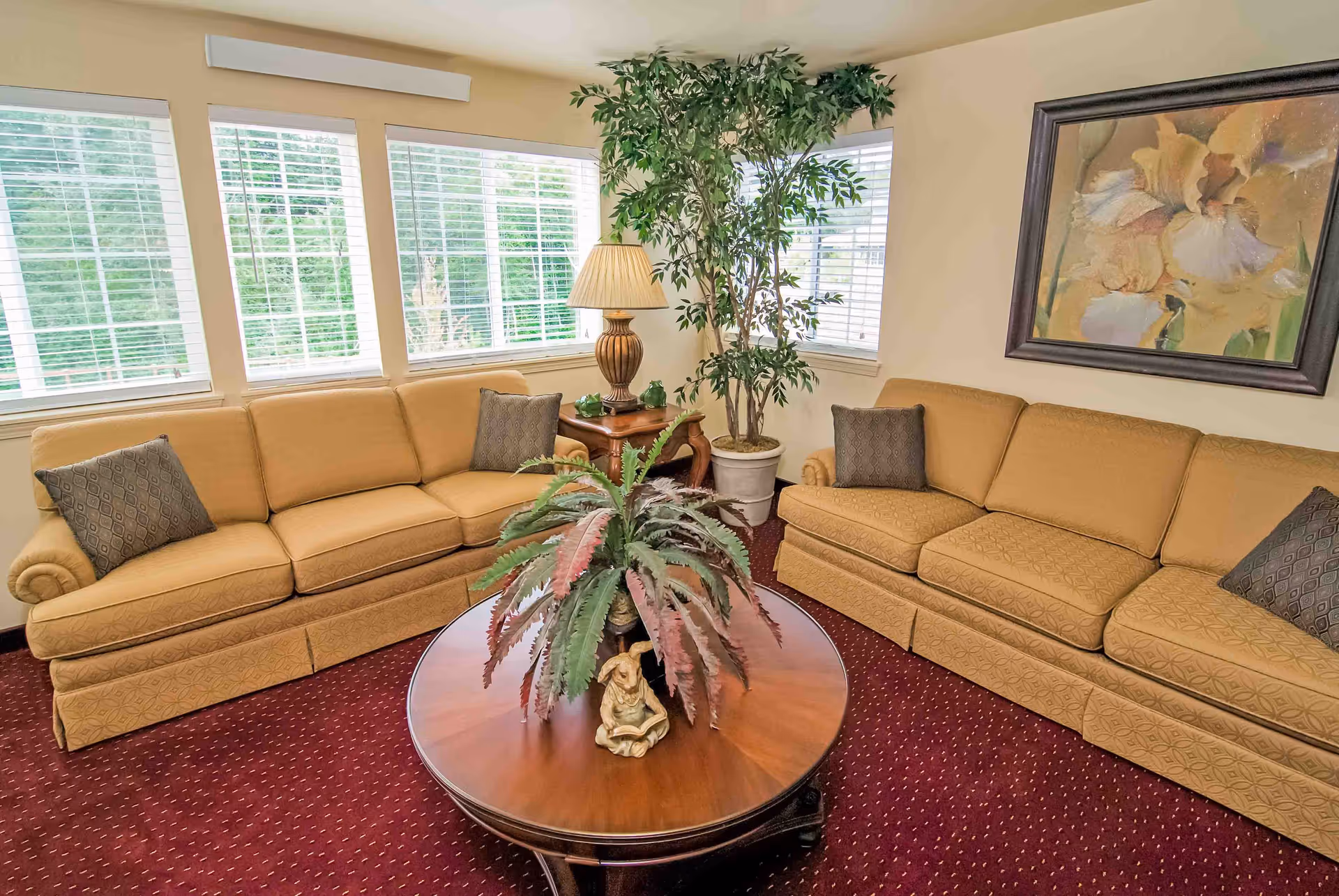 A cozy sitting area with two beige sofas facing a round wooden coffee table, potted plants, a table lamp, and large windows with blinds.
