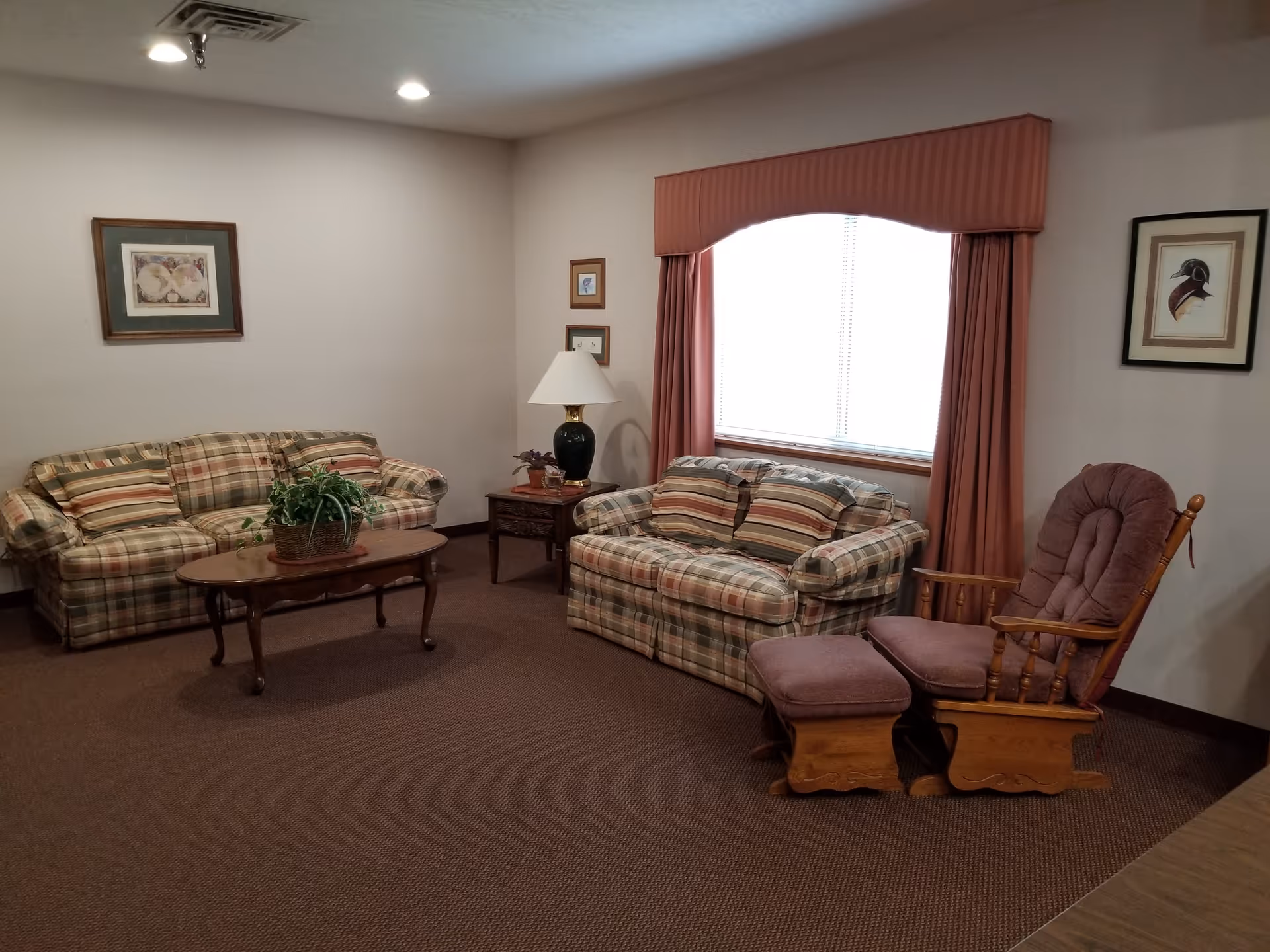 A cozy communal living room with plaid sofas, a coffee table, side tables with a lamp, and a cushioned wooden rocking chair.