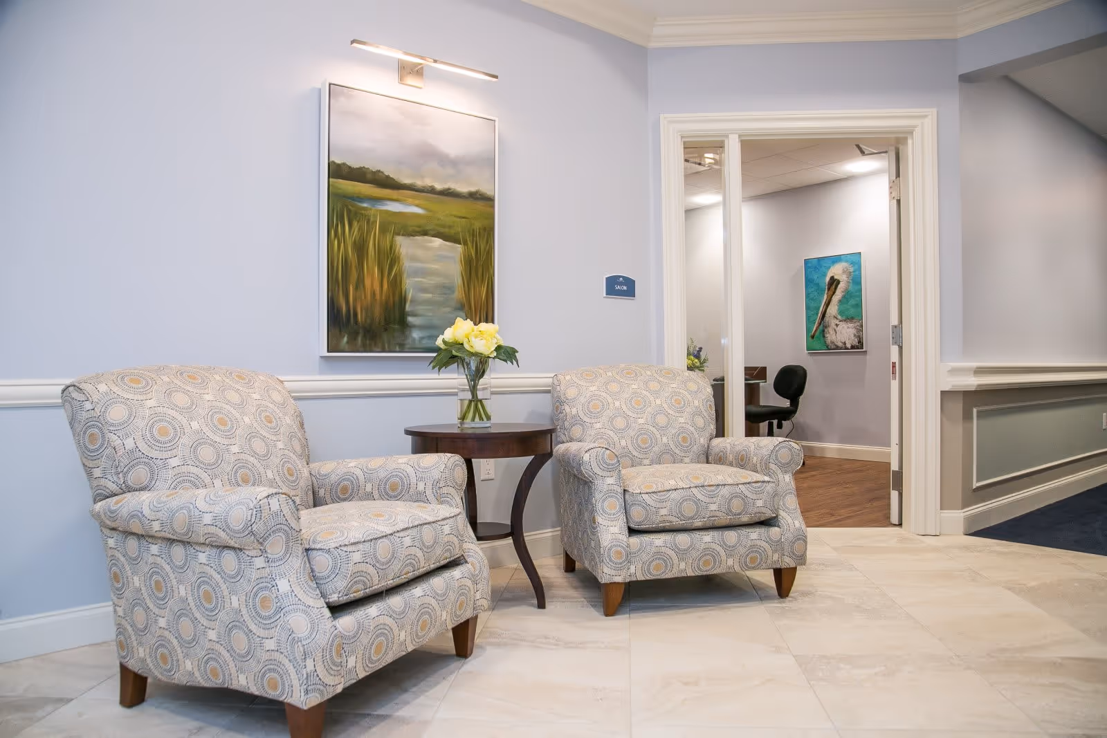 A cozy seating area with two patterned armchairs and a small round wooden table with a vase of yellow flowers between them. The walls are painted light blue with white trim, and a landscape painting hangs above the chairs. An open door leads to another room with a chair and a painting of a pelican on the wall.