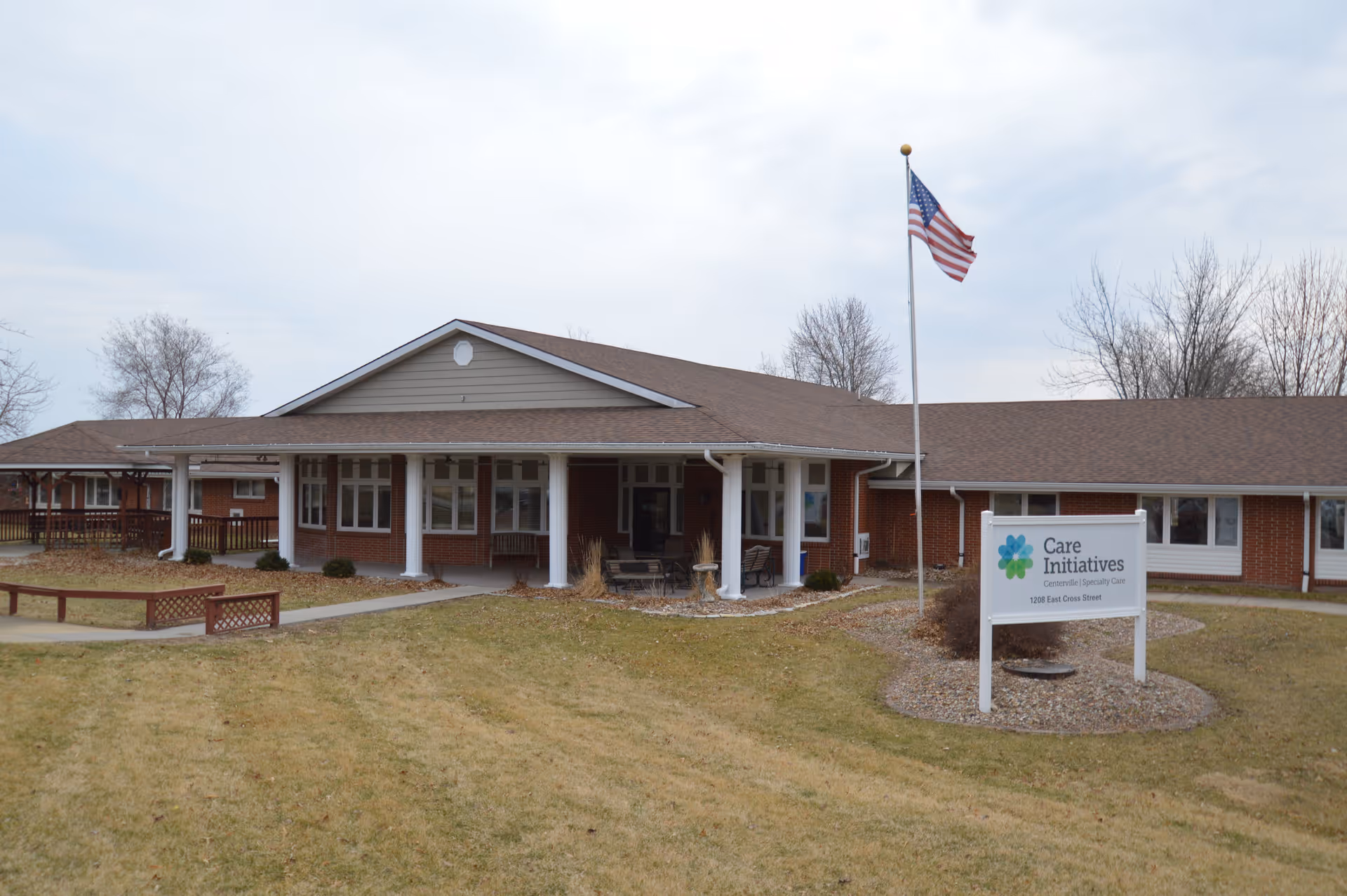 Front exterior of a single-story brick senior care facility with a covered entrance, American flag on a flagpole, and a 'Care Initiatives' sign on the lawn.