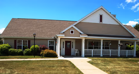 Single-story building with beige siding and a brown shingled roof, featuring a covered porch with white railings and columns. There is a concrete walkway leading to the entrance, surrounded by grass and bushes under a blue sky with some clouds.