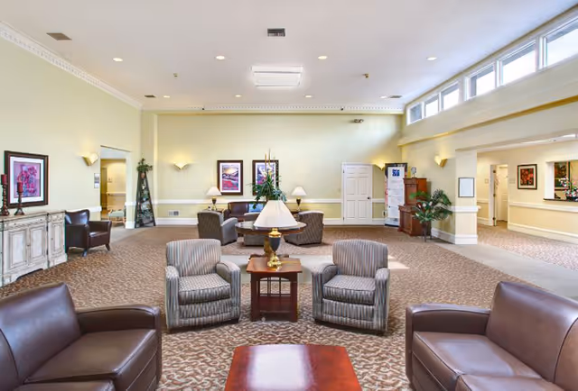 A spacious and well-lit living room area with beige walls and carpeted floor. The room features multiple seating arrangements including brown leather sofas and striped armchairs around wooden coffee tables. There are framed artworks on the walls, table lamps, and plants adding to the decor. Large windows near the ceiling allow natural light to brighten the space.