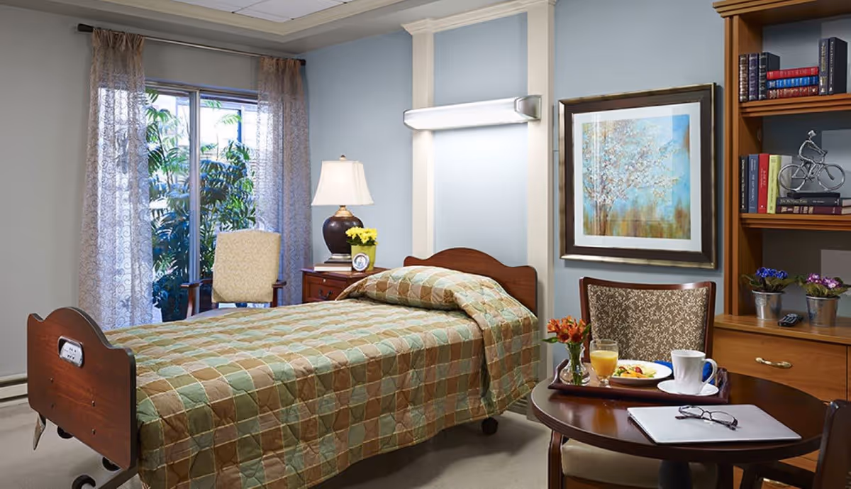 A neatly arranged bedroom in a senior living facility featuring a single bed with a checkered bedspread in shades of green and brown. Next to the bed is a wooden nightstand with a lamp, a small framed photo, and a yellow flower pot. A window with sheer curtains reveals greenery outside. A round wooden table holds a tray with a breakfast plate, a glass of orange juice, a cup of coffee, a small vase with flowers, and a pair of glasses resting on a closed laptop. A wooden bookshelf with books and decorative items is visible on the right wall, along with a framed painting of a tree.