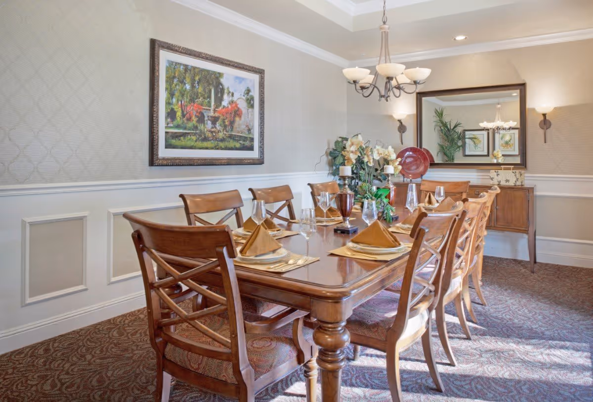 A formal dining room with a long wooden table set for eight people. The table is decorated with folded brown napkins, glassware, and a centerpiece of flowers and candles. The room has beige patterned wallpaper, a large framed painting on one wall, a large mirror on another wall, and a chandelier hanging from the ceiling. There is a wooden sideboard beneath the mirror with decorative items on it.