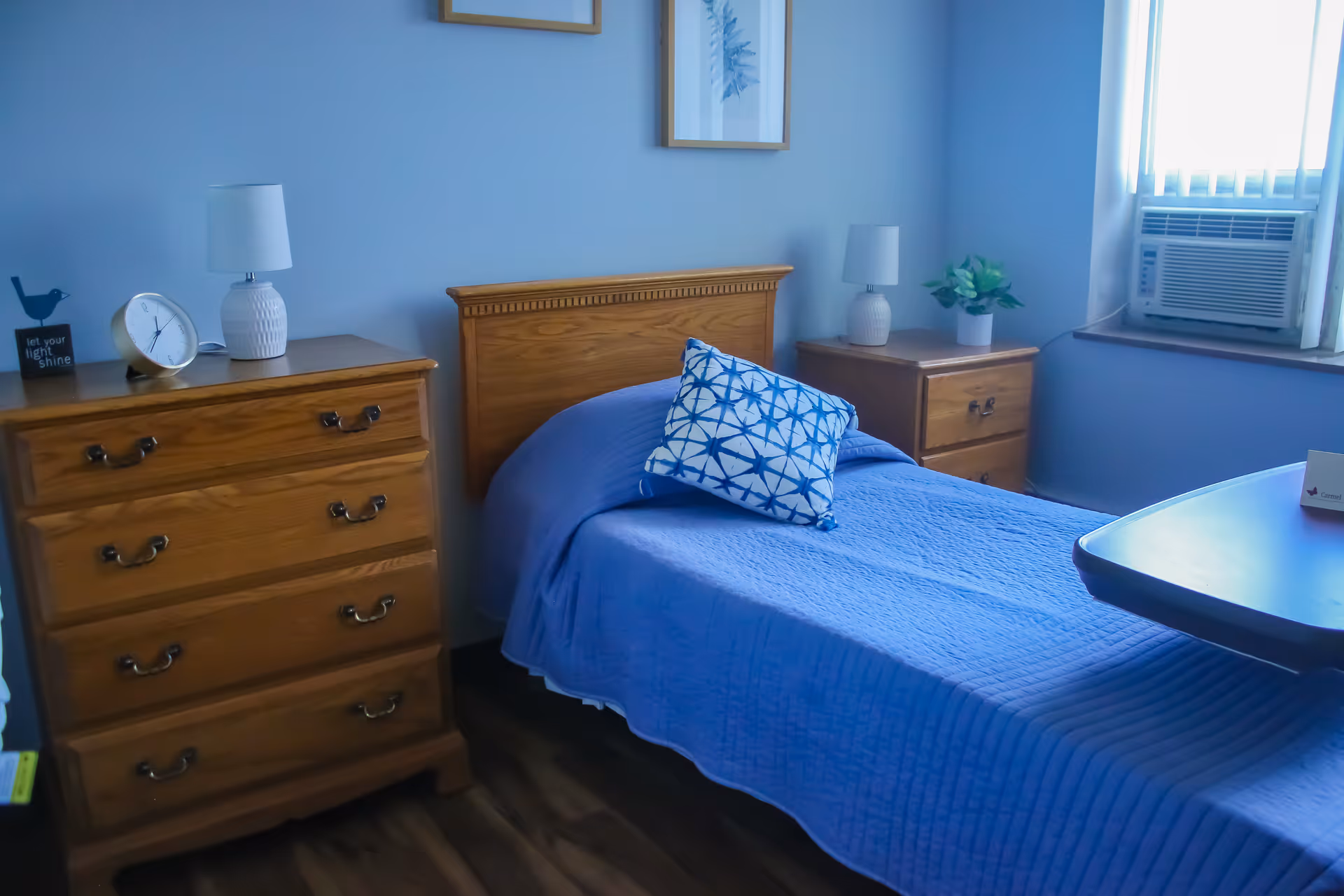 A neatly made single bed with a blue quilt and a decorative blue and white pillow in a room with light blue walls. There is a wooden headboard, a wooden dresser with a white lamp and clock, and a wooden nightstand with a white lamp and a small plant. A window with vertical blinds and an air conditioning unit is visible on the right side of the room.