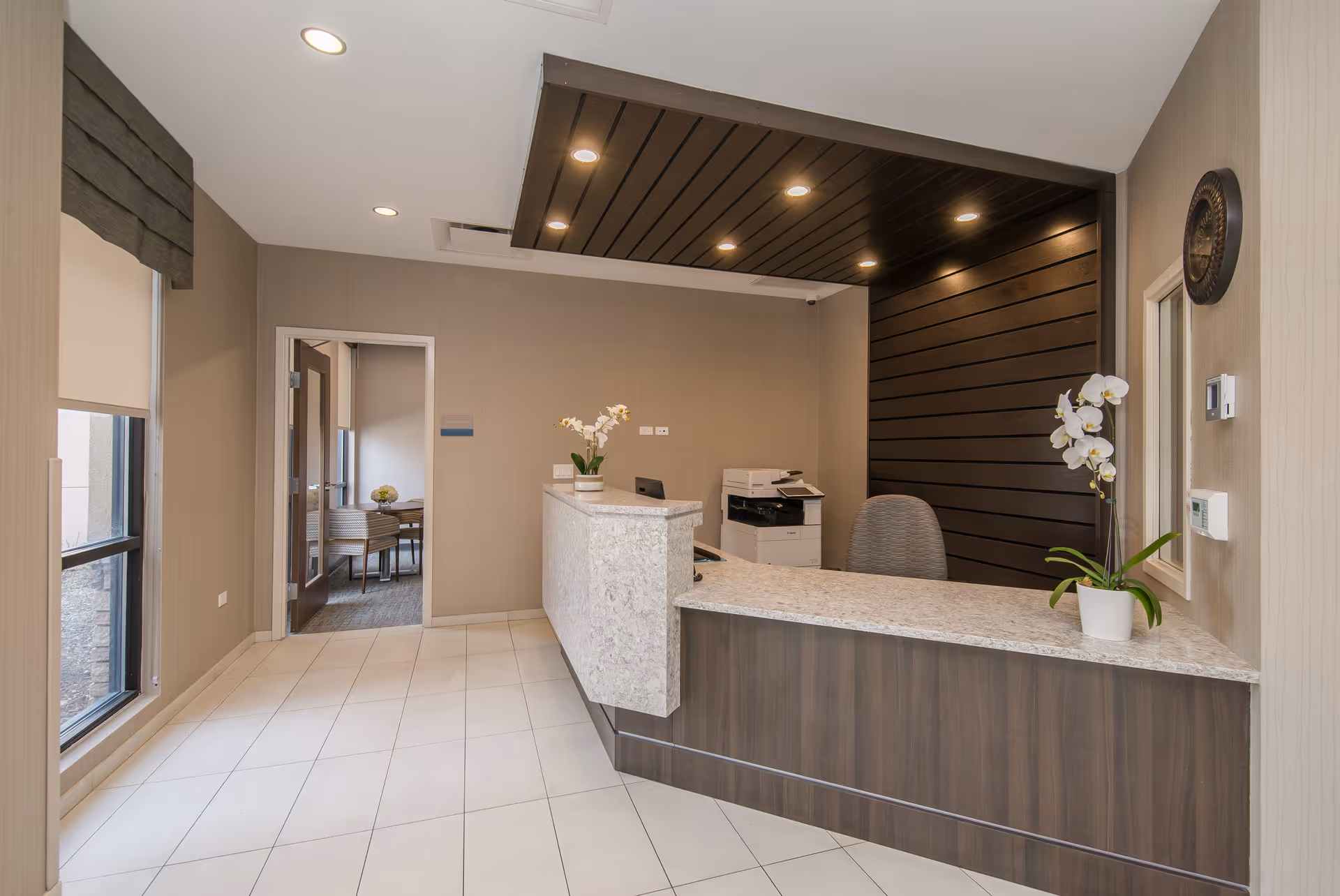 Reception area with a modern desk featuring a granite countertop and dark wood paneling behind it. There is a potted orchid plant on the desk, a chair, and a printer in the corner. The room has beige walls, tiled floor, recessed lighting, and a window with a shade on the left side. An open doorway leads to another room with a table and chairs.
